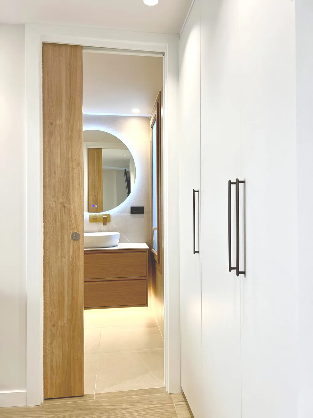 Pasillo con puerta de madera y baño con espejo redondo iluminado · Ana Uría Interiorismo